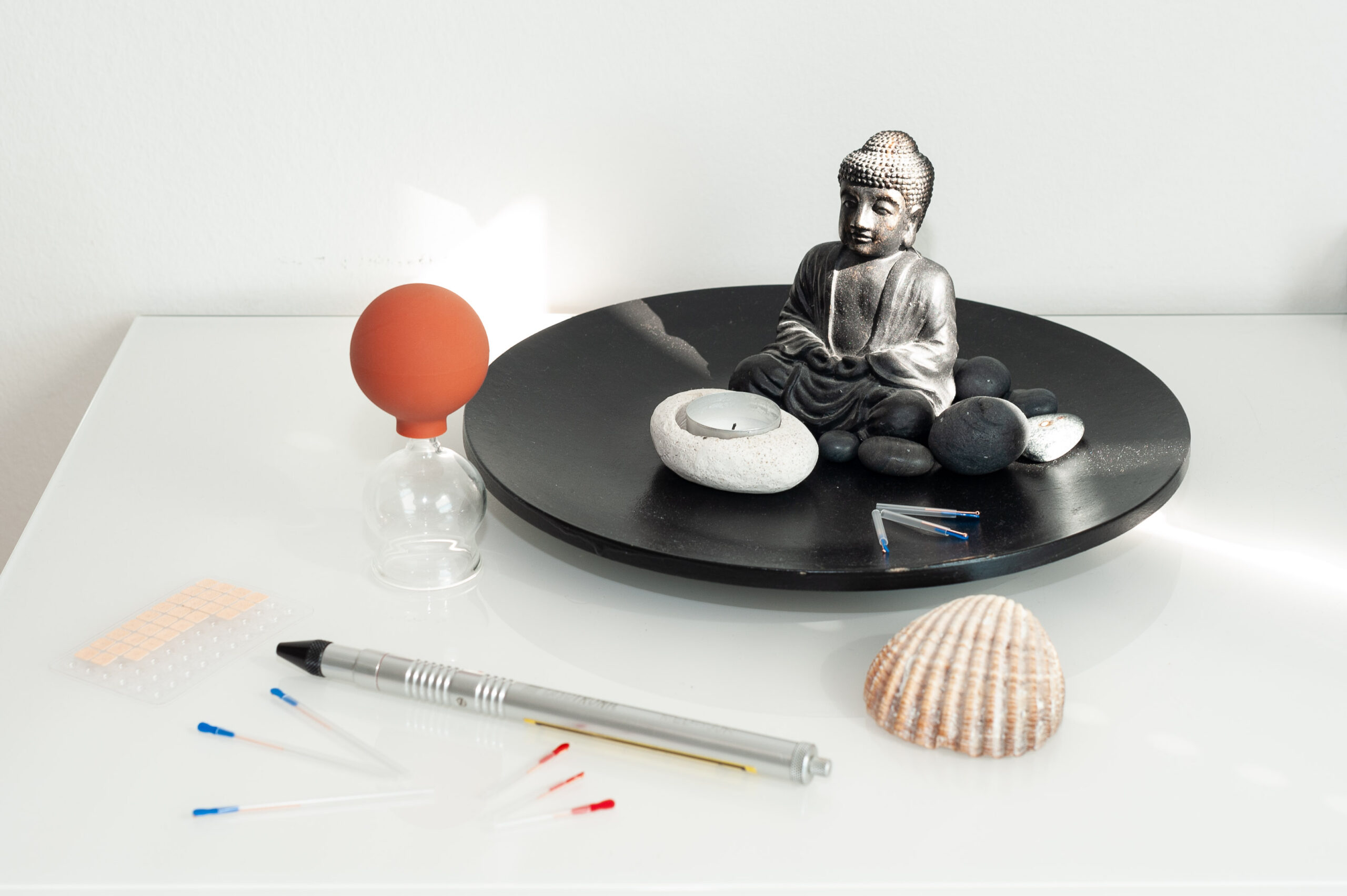 Eine kleine Buddastatue auf einem Teller, ein Schröpfglas, eine Muschel, eine Kerze, Akupunkturnadeln und vieles weiteres arrangiert in der Praxis von Dr. Helga Barilich.