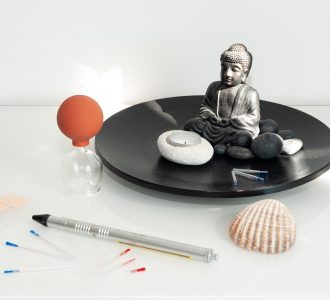 Eine kleine Buddastatue auf einem Teller, ein Schröpfglas, eine Muschel, eine Kerze, Akupunkturnadeln und vieles weiteres arrangiert in der Praxis von Dr. Helga Barilich.